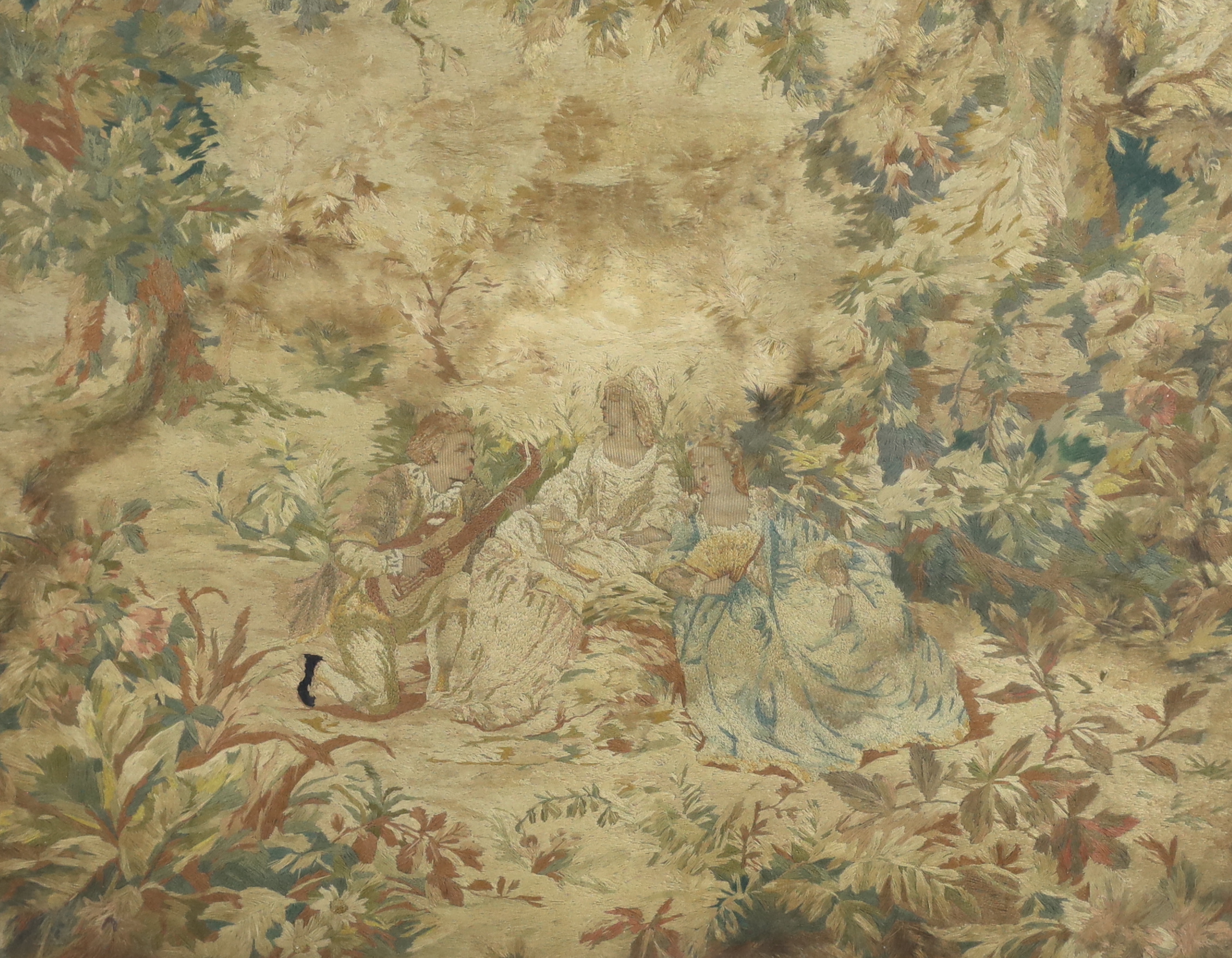 A mid 19th century framed ornate silk embroidery panel of two seated ladies being serenaded by a young musician in a luscious wooded landscape, 64cm wide x 49cm high